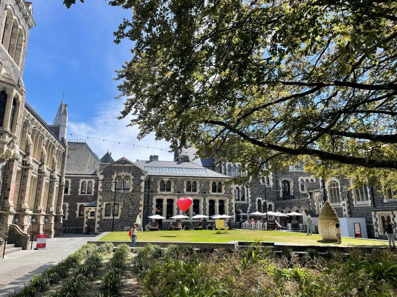 "Grüner Innenhof der Universität. Ein großer Baum spendet Schatten über eine Fläche an der kleiner Pflanzen wachsen. Im Hintergrund stehen Tische mit kleinen Stühlen und Schirmen an denen vereinzelt Menschen im Schatten sitzen. Davor ist eine kleine begrünte Fläche, die von der Sonne bestrahlt wird. Ein roter herzförmiger Luftballon steigt über den Tischen auf."