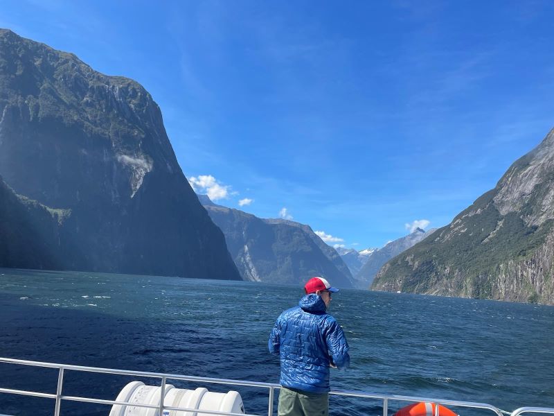 "Manu steht in seiner blauen Jacke auf dem Boot. Er schaut auf das blaue Wasser. Im Hintergrund ist eine Berglandschaft und blauer Himmel zu erkennen."