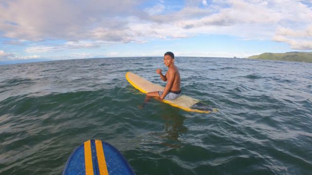 "Chad im Meer, wie er auf seinem gelben Surfbrett sitzt, lächelt und seine Faust fröhlich vor sich hält."