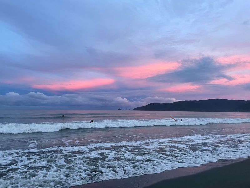 "Die Abenddämmerung bricht ein über dem North Shore Beach in Baler. Eine Wolkengruppe hängt über den Bergen, darüber rosarote Wolken. Im Meer bricht eine Welle. Einige Surfer sind im Wasser."