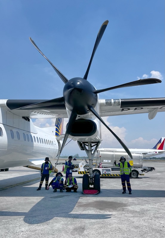 "Unter dem riesigen Propeller des Flugzeugs stehen fünf Flugzeugangestellte. Zwei kauern auf der einen Seite am Boden und posieren mit Handgesten. Daneben und der hinter stehen auch zwei die lächeln. Auf der anderen Seite gesteht noch einer und hält zwei Finger in die Luft zum Victory-Zeichen"