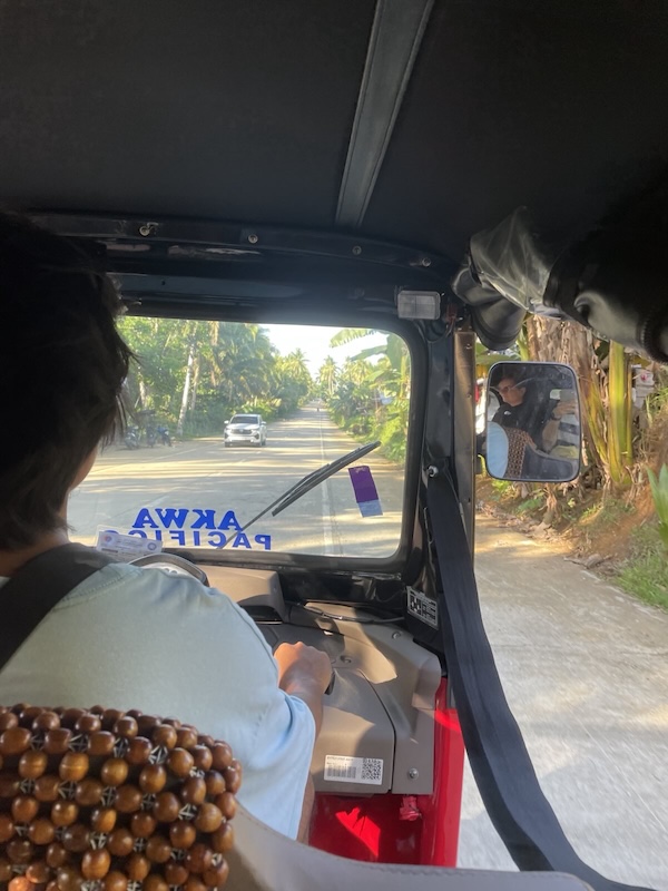 "Blick vom Rücksitz des Tuktuks. Wir schauen David über die Schulter. Auf der Windschutzscheibe steht spiegelverkehr Akwa Pacifico. Hinter der Windschutzscheibe kommt uns ein weißes Auto entgegen. Wir sind umgeben von Kokosnusspalmen."