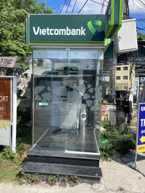 "Eine gläserne Kabine mit zwei Stufen. Darin befindet sich ein Bankomat, darauf steht auf waldgrünen Hintergrund in weißer Schrift: Vietcombank"