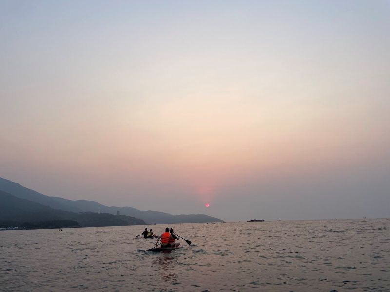 Die rote Sonne geht auf. Der Himmel wird langsam hell, darunter erheben sich blaue Berge und davor ist das Meer. Zwei Paddelbretter schwimmen im Wasser mit jeweils zwei Personen an Bord.