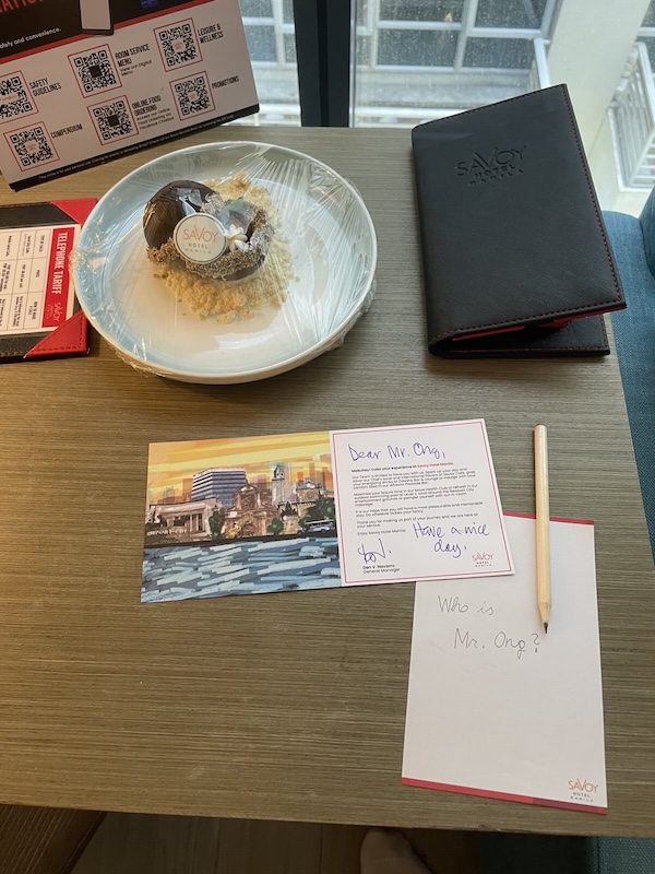 "Am Tisch steht ein Dessert auf einem Teller, der mit Frischaltefolie überzogen wurde. Darunter liegt eine Karte."
