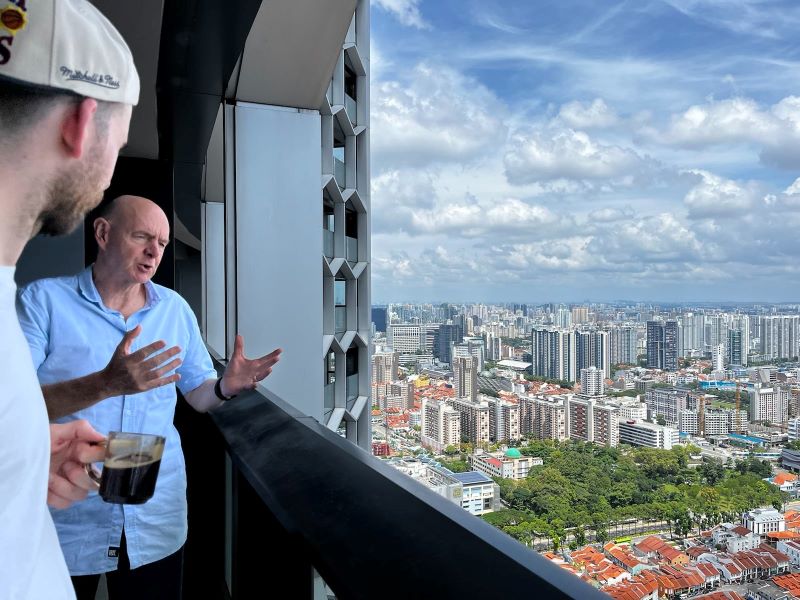 Ich bin im Profil sichtbar, wie ich ein Glas mit Kaffee halte. Neben mir steht David in blauem Hemd. David hat eine Glatze und hält seine Hände vor sich ausgestreckt. Vom Balkon aus ist der Himmel und die Skyline von Singapur zu sehen. Darunter die roten Dächer von Kampong Glam.