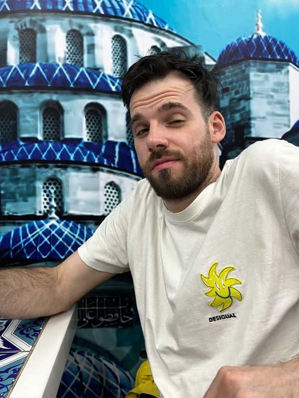 Ich sitze zu Tisch, hinter mir sieht man ein Bild einer Moschee mit blauem Dach. Ich trage ein weißes T-Shirt mit einem Sonnen-Logo auf der Brust unter dem der Schriftzug "Desigual" steht. Ich verziehe das Gesicht und hab die Augen zugeknfeift.