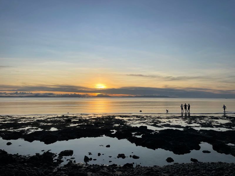 "Der Sonnenuntergang beim Secret Beach von Tangbo. Es herrscht Ebbe, also erkennt man schroffe Felsen vorm Meer. Der Umriss von einige Personen und ein Hund sind im seichten Wasser erkennbar."