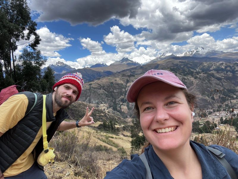 Ein Landschaftsfoto. Vor einer Berglandschaft und einem stark bewölkten Himmel stehen Sophia und ich. Sophia trägt eine rote Kappe, ein blaues Hemd und lächelt. Ich trage eine Wollhaube mit rot-weißem Muster, eine Weste und ein gelbes T-Shirt.