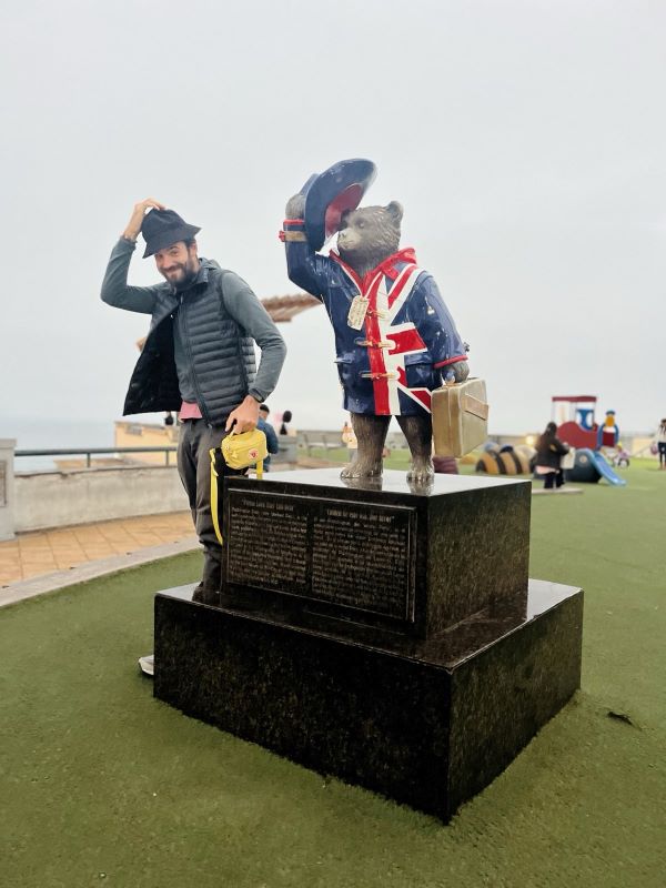 Die Statue von einem Bär auf einem Marmorpodest. Der Bär trägt einen Mantel mit britischer Flagge und zückt mit rechter Hand seinen Hut. Ich stehe daneben und imitiere die Pose, indem ich ebenfalls meinen Hut zücke.