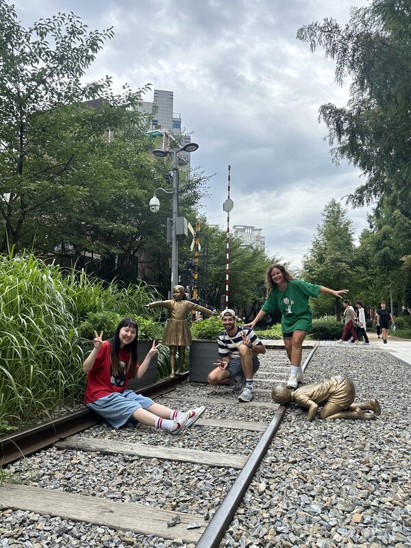 Mitten durch einen grünen Park verlaufen Bahnschienen. Sophia und ich posieren auf den Bahngleisen mit unserer Freundin Sani neben zwei Bronzestatuen von Kindern.